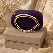 Purple sculptural clutch with gold-toned loop handle, placed on gift box.