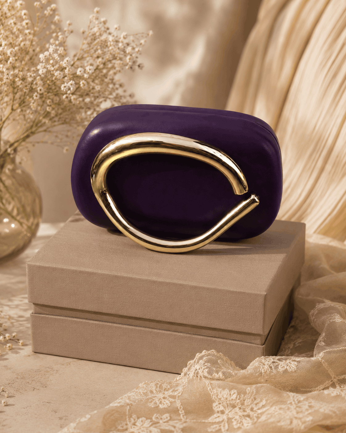 Purple sculptural clutch with gold-toned loop handle, placed on gift box.
