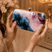 Alora clutch close-up held with hands: gold-tone frame, blue and pink marble print.