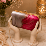 Alora clutch held by hands: gold-tone frame, pink and red marble print, chain strap.