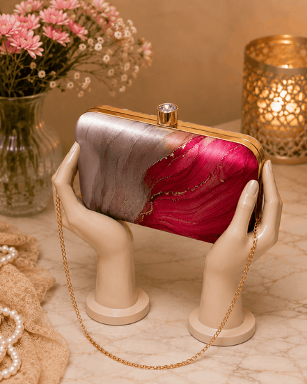 Alora clutch held by hands: gold-tone frame, pink and red marble print, chain strap.