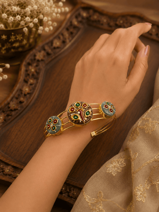 Gold-tone cuff bracelet worn on wrist, featuring colorful peacock and round enamel charms
