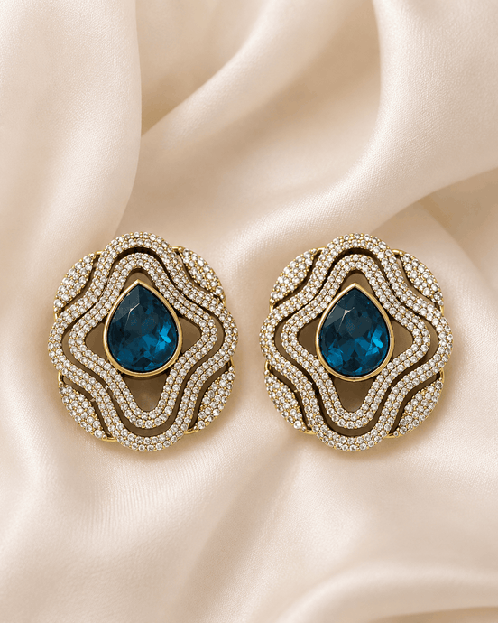 Brass-tone statement stud earrings with blue teardrop center and layered crystal trim